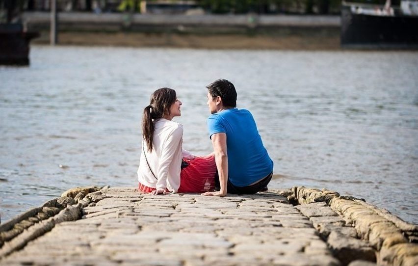 Couple smile at each other sitting by the Thames on their Southbank pre-wedding shoot