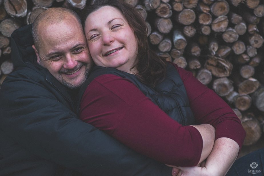 A couple hugging and laughing in front of a pile of stacked logs