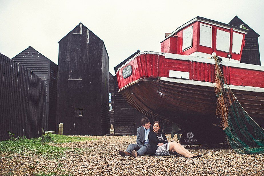 An engaged couple sitting under a fishing boat surrounded by old black fishing huts on their Hastings pre wedding shoot