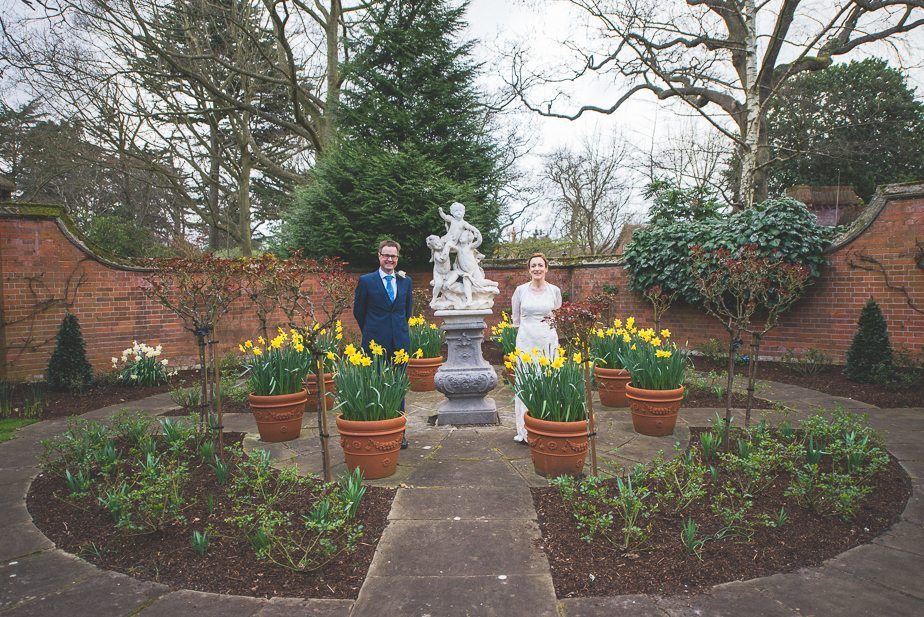 A bride and groom standing by the daffodil fountain at Warren House