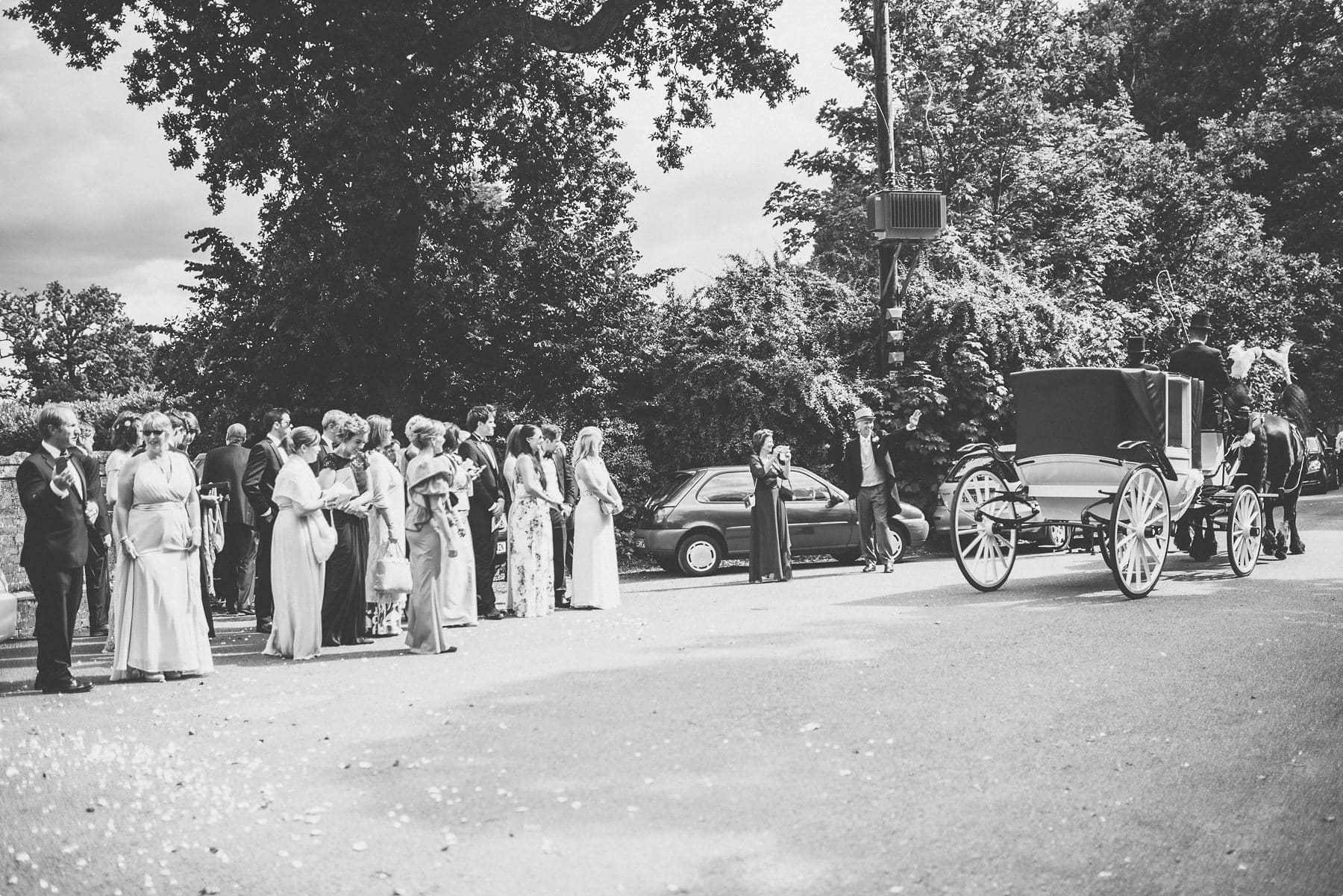 Wedding guests waving off the bride and groom in their horse drawn carriage bound for their Coworth Park barn summer wedding photography