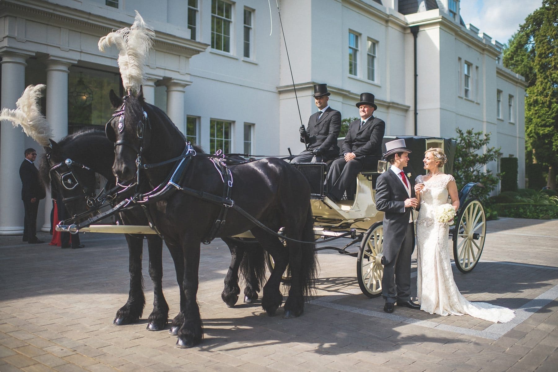 Bride and groom smiling at each other standing by their horse drawn carriage at their Coworth Park Barn summer wedding