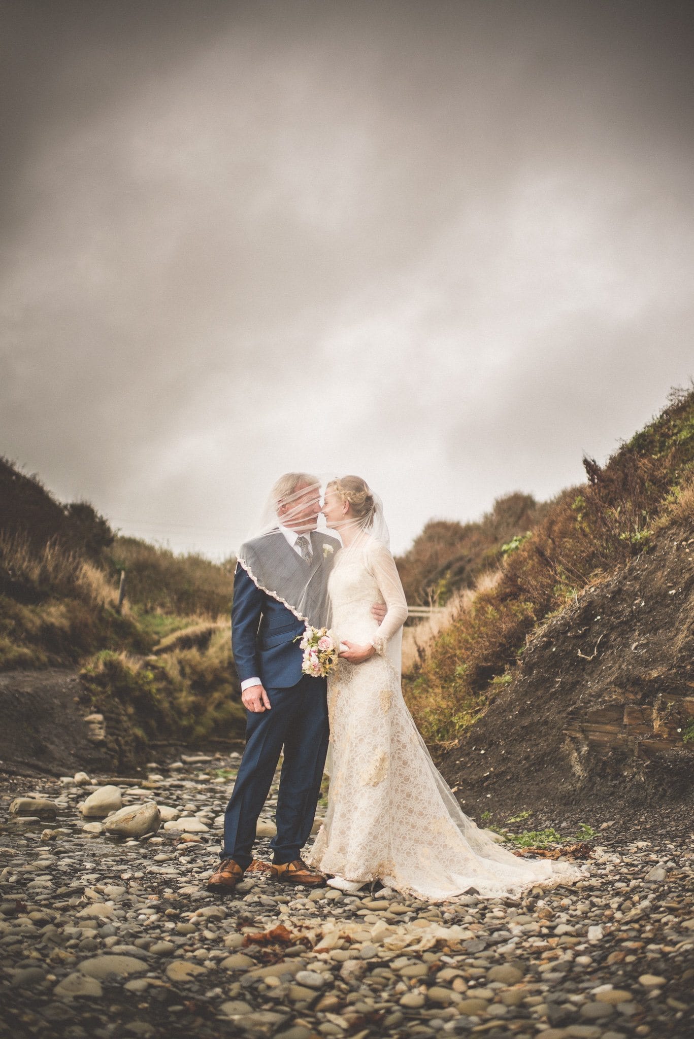 Bride and Groom stand together in Kimmeridge Bay with her veil blowing around them