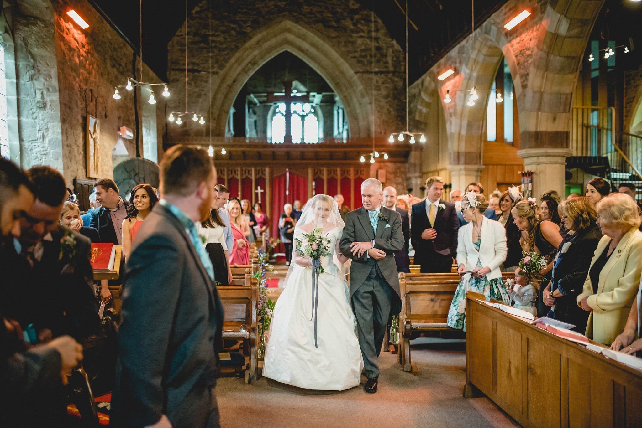 Hatty walks down the aisle on her father's arm, holding her bouquet with a trailing grey ribbon