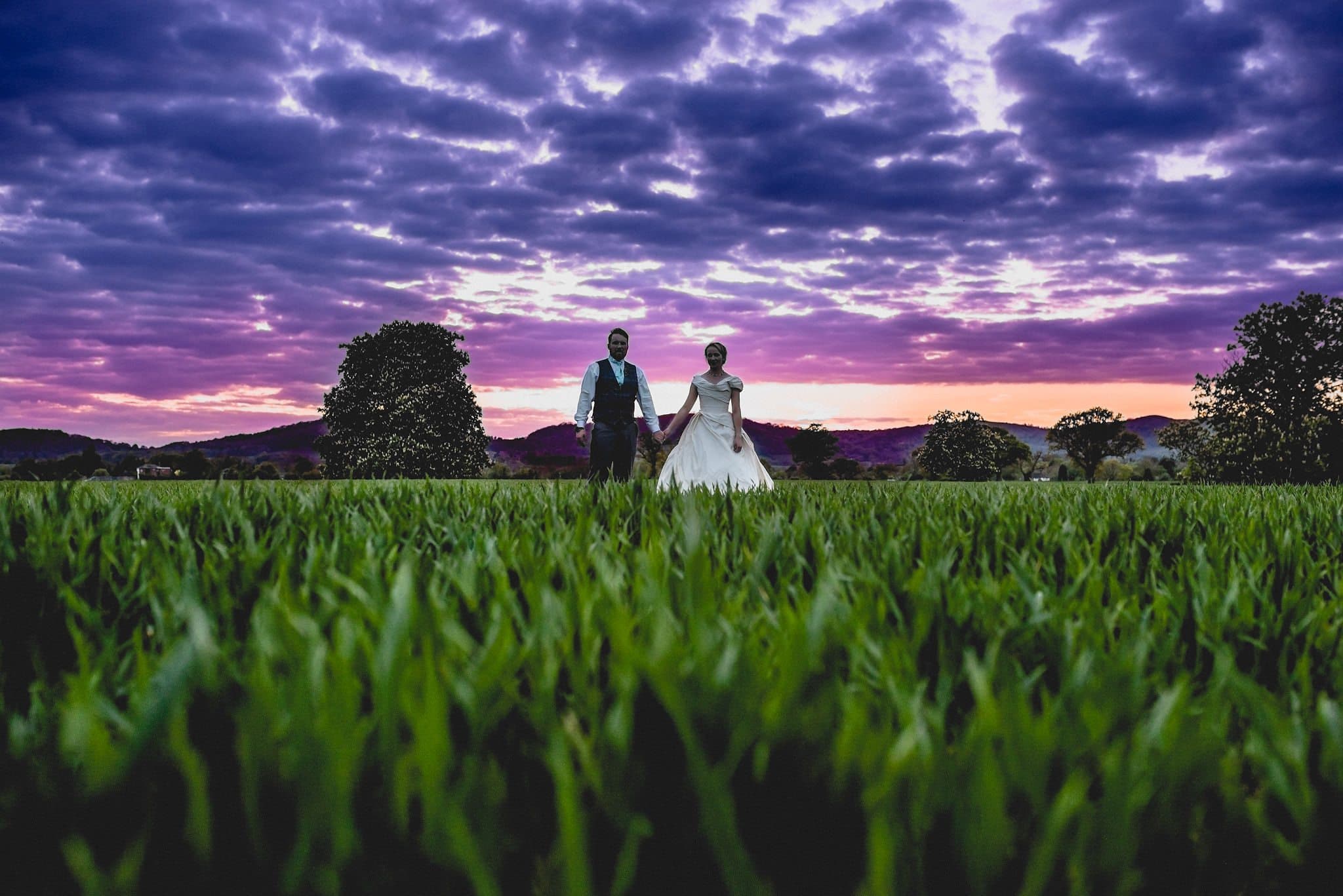 Bride and groom stand in the long grass at sunset overlooking the malvern hills