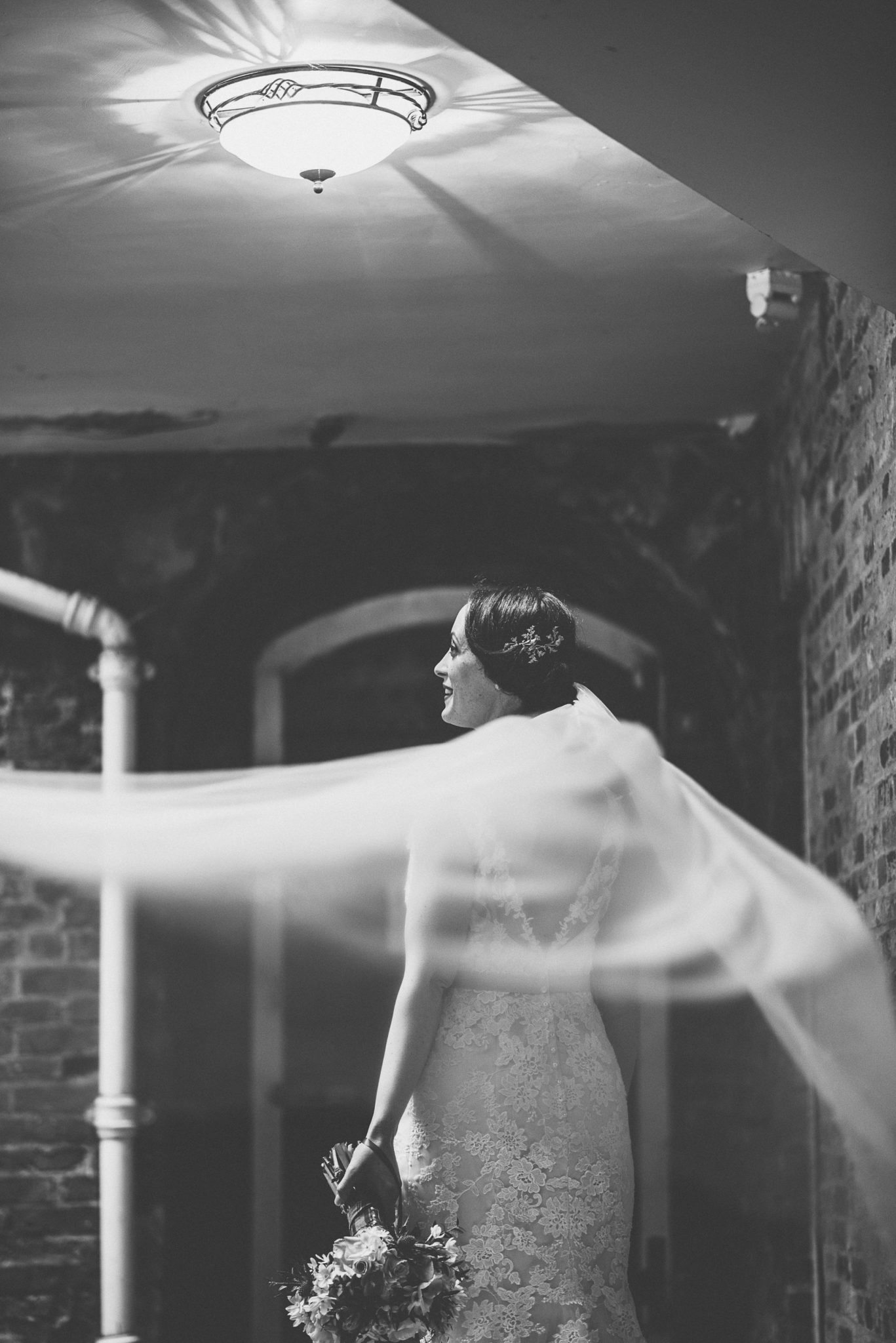 Black and White photo of the bride standing below a light with her bridal veil blowing in the wind