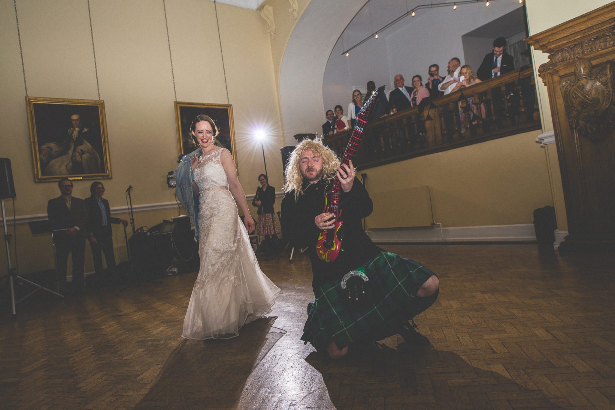 Scottish Groom in a long blond wig plays an inflatable guitar while his bride laughs