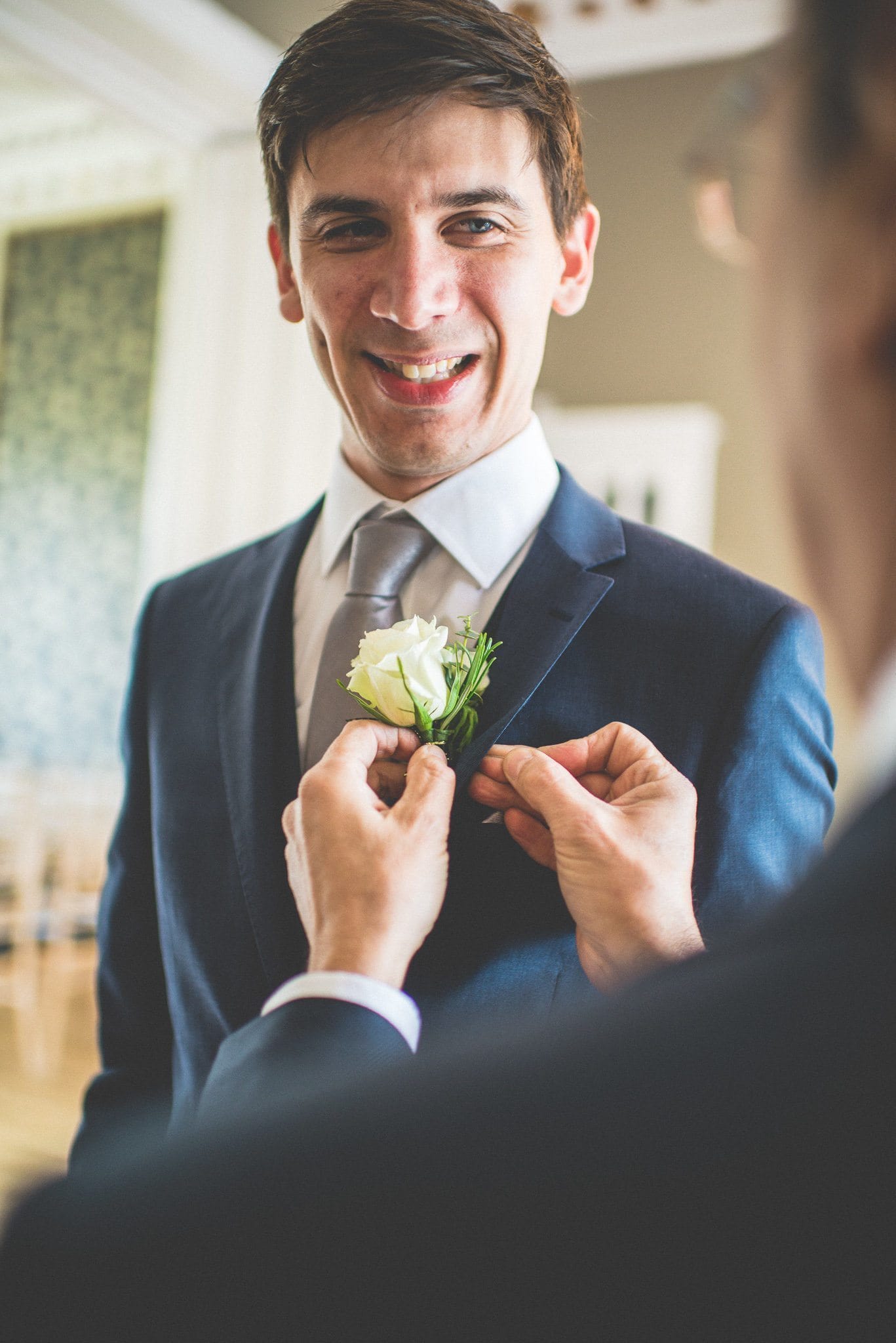 A groomsman fastens Andy's buttonhole to his navy suit