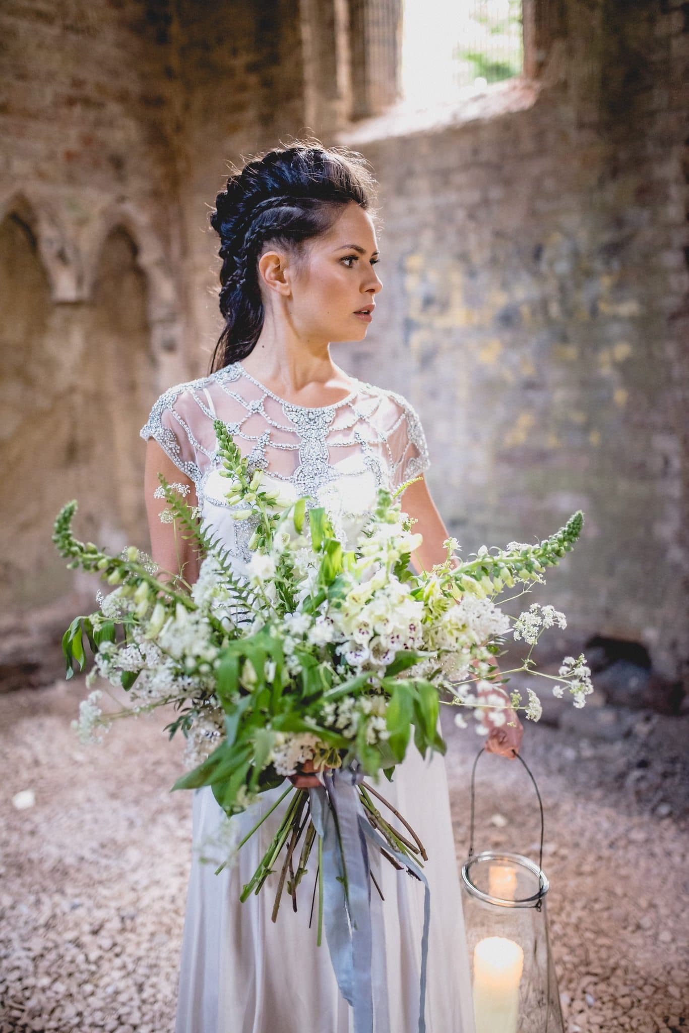 Bride with elaborately braided hair carries large bouquet and storm lantern with candle