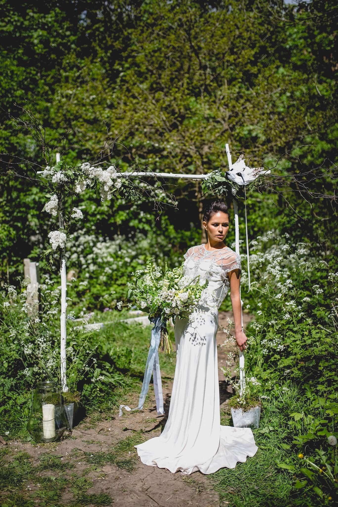 Bride leans against wooden arch decorated with flowers, with feather fox mask in top right corner