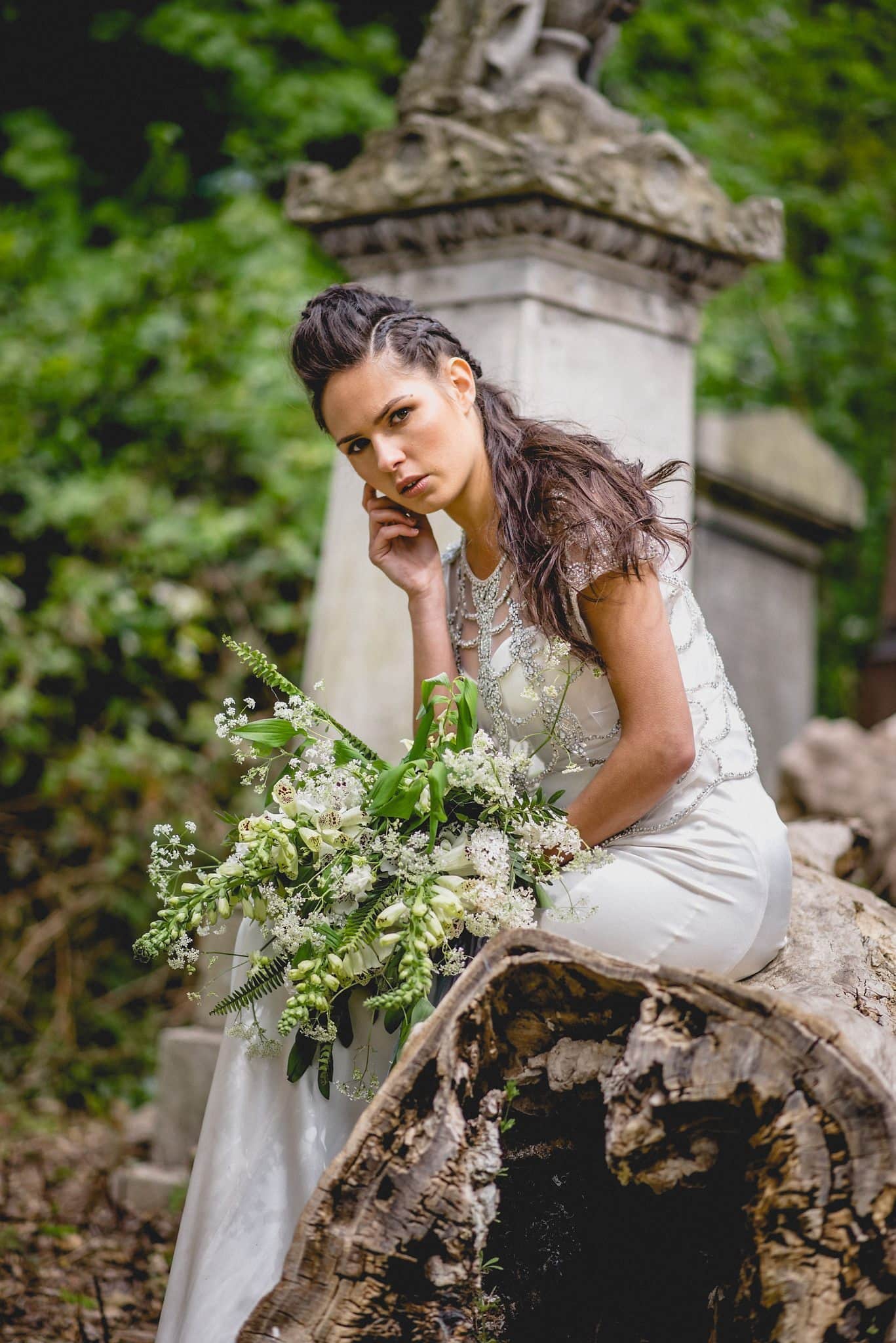 Bride sits on fallen tree, leaning forward and resting her chin on her hand