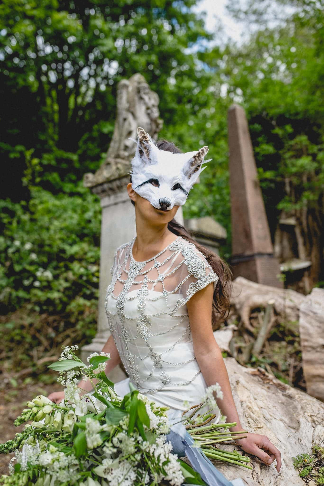 Bride seated on fallen tree wearing feather fox mask among the tombs in Abney Park Cemetery