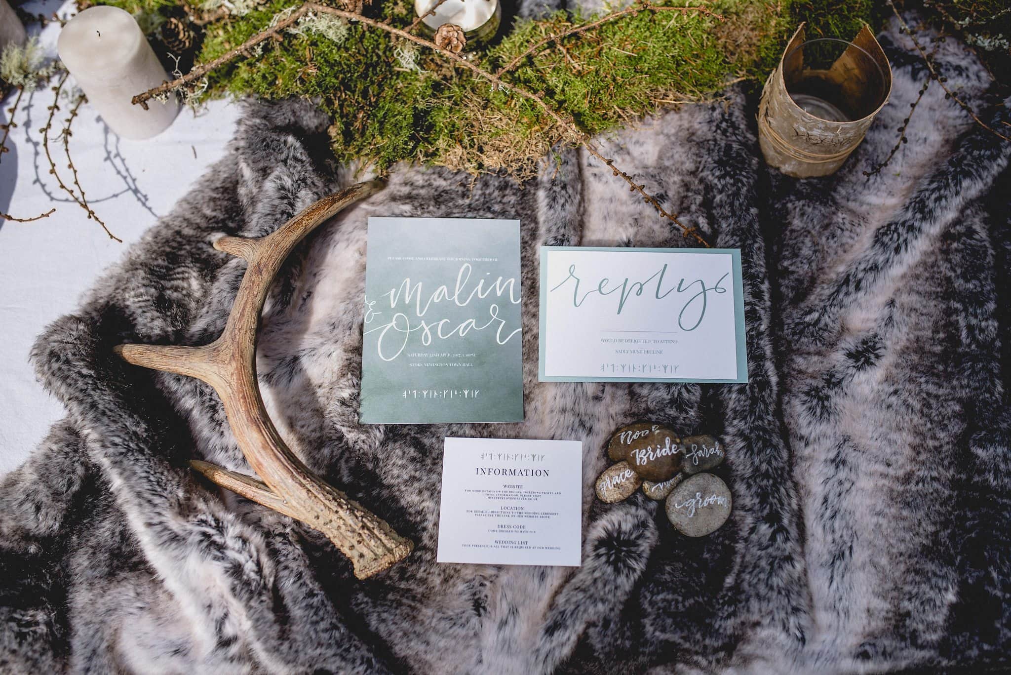 Flatlay of sage green stationery on grey fur throw with antler, moss and pebbles