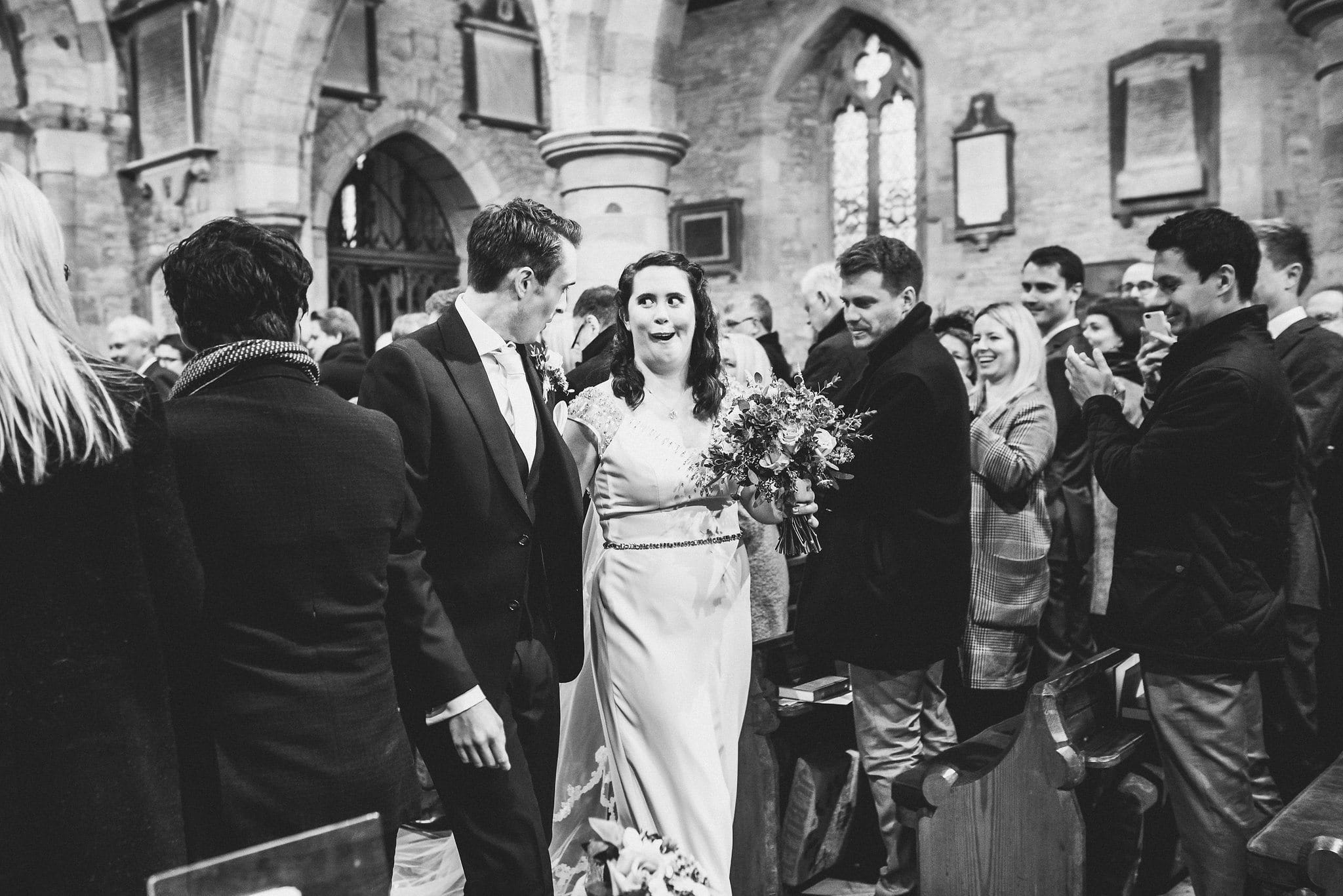 Black and white shot of bride and groom walking back down aisle. Bride pulling funny face