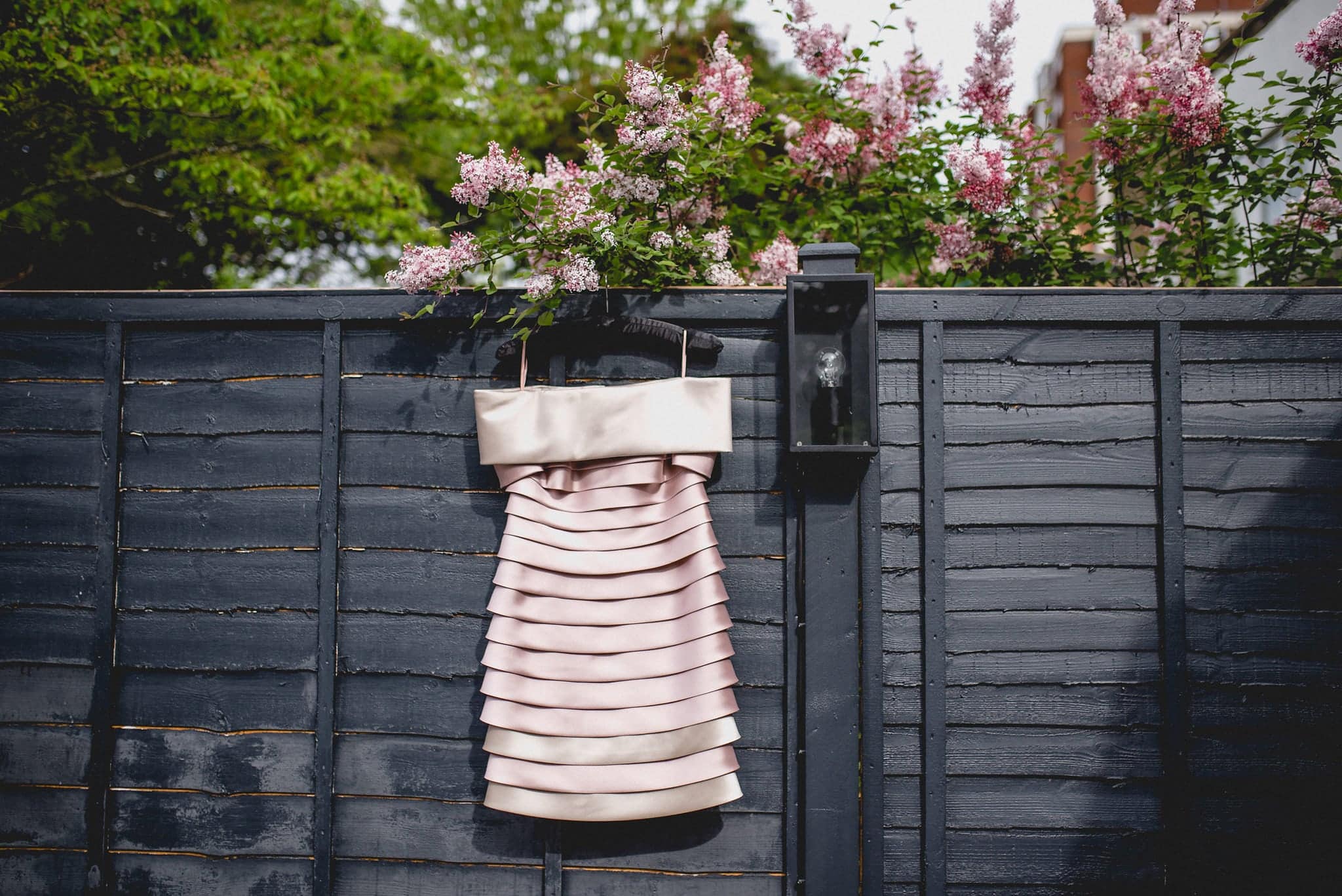 Alternative ruffled pink wedding dress hanging on black fence with lilac in the background