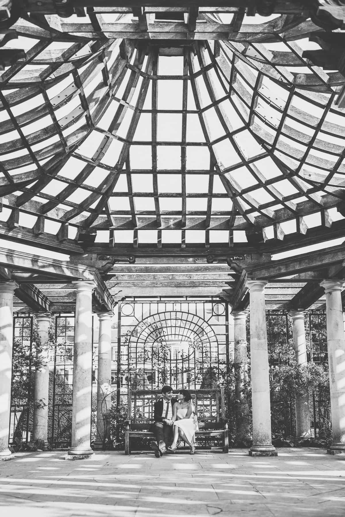 The stunning beam roof of the Hampstead Pergola surrounds the bride and groom cuddling on a bench