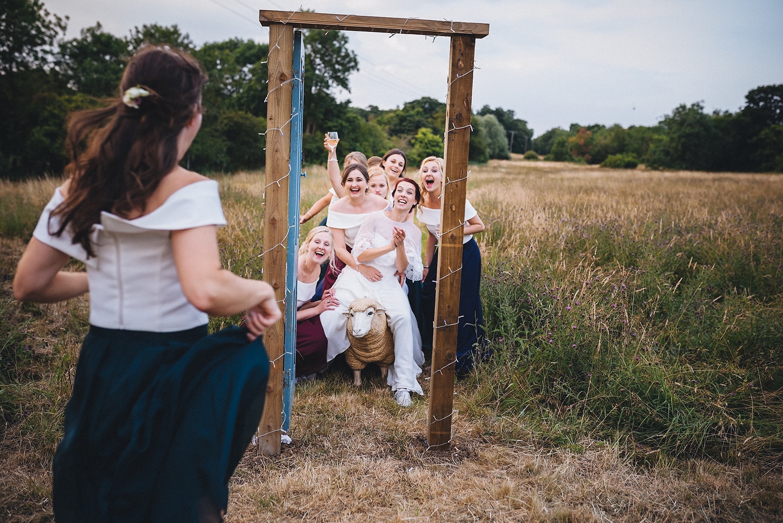 Bride laughing sitting on a sheep framed by a doorframe in a field surrounded by her bridesmaids