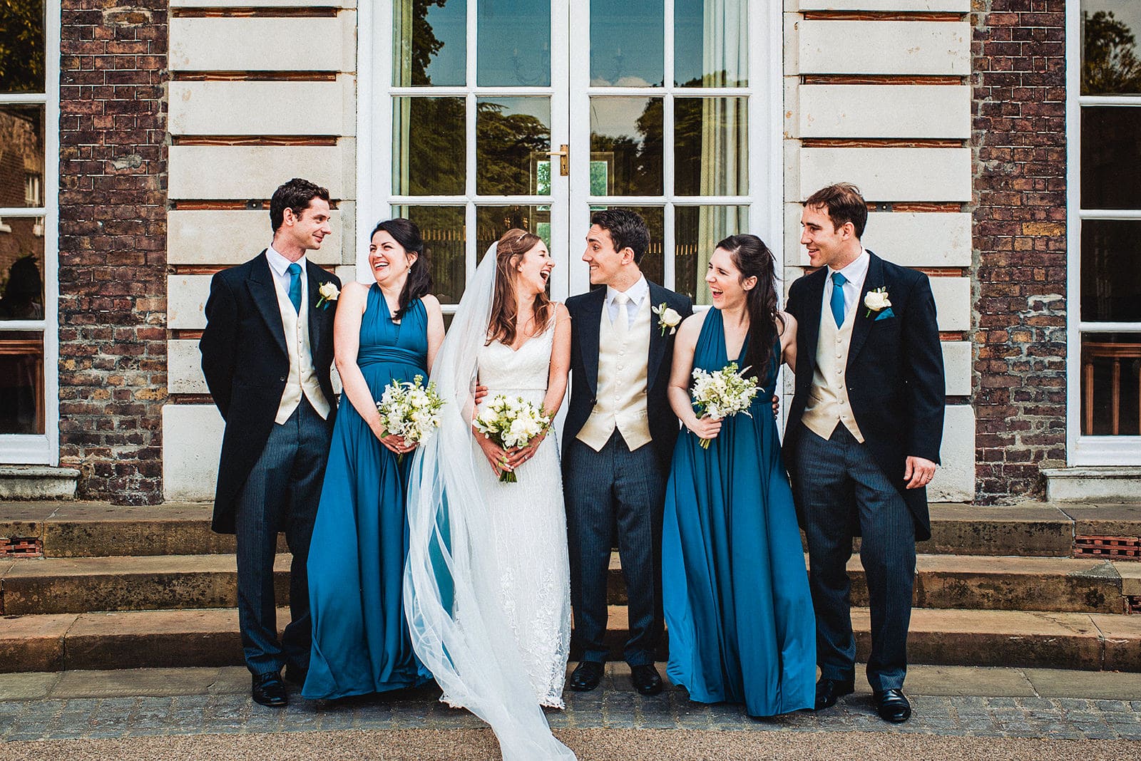 Bride and groom, teal bridesmaids and groomsmen laugh outside the Hurlingham Club