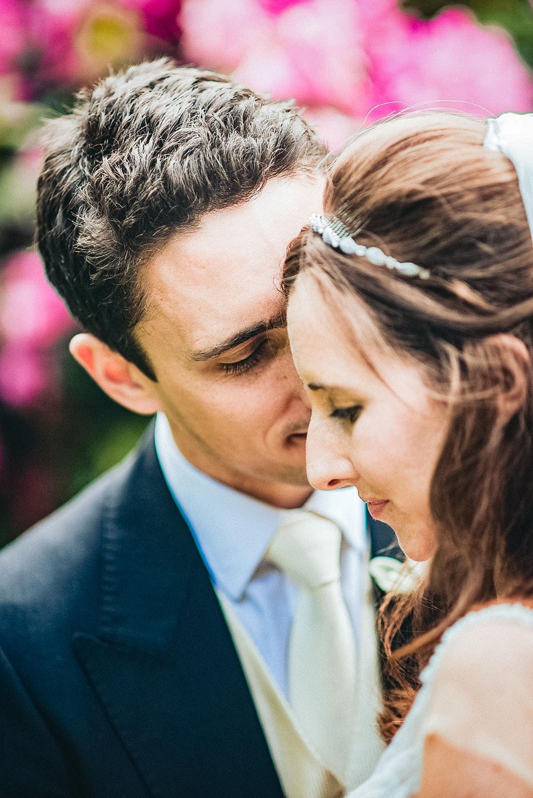 Groom kisses the bride's cheek with pink flowers at the hurlingham Club behind them