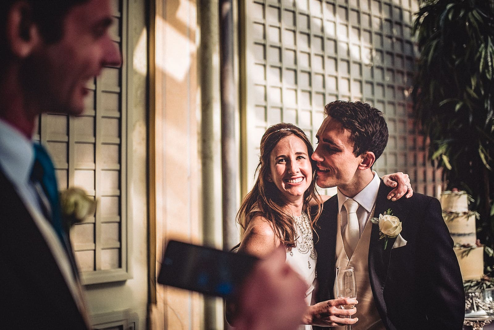 Groom hugs his bride while a wedding guests take a photo of them