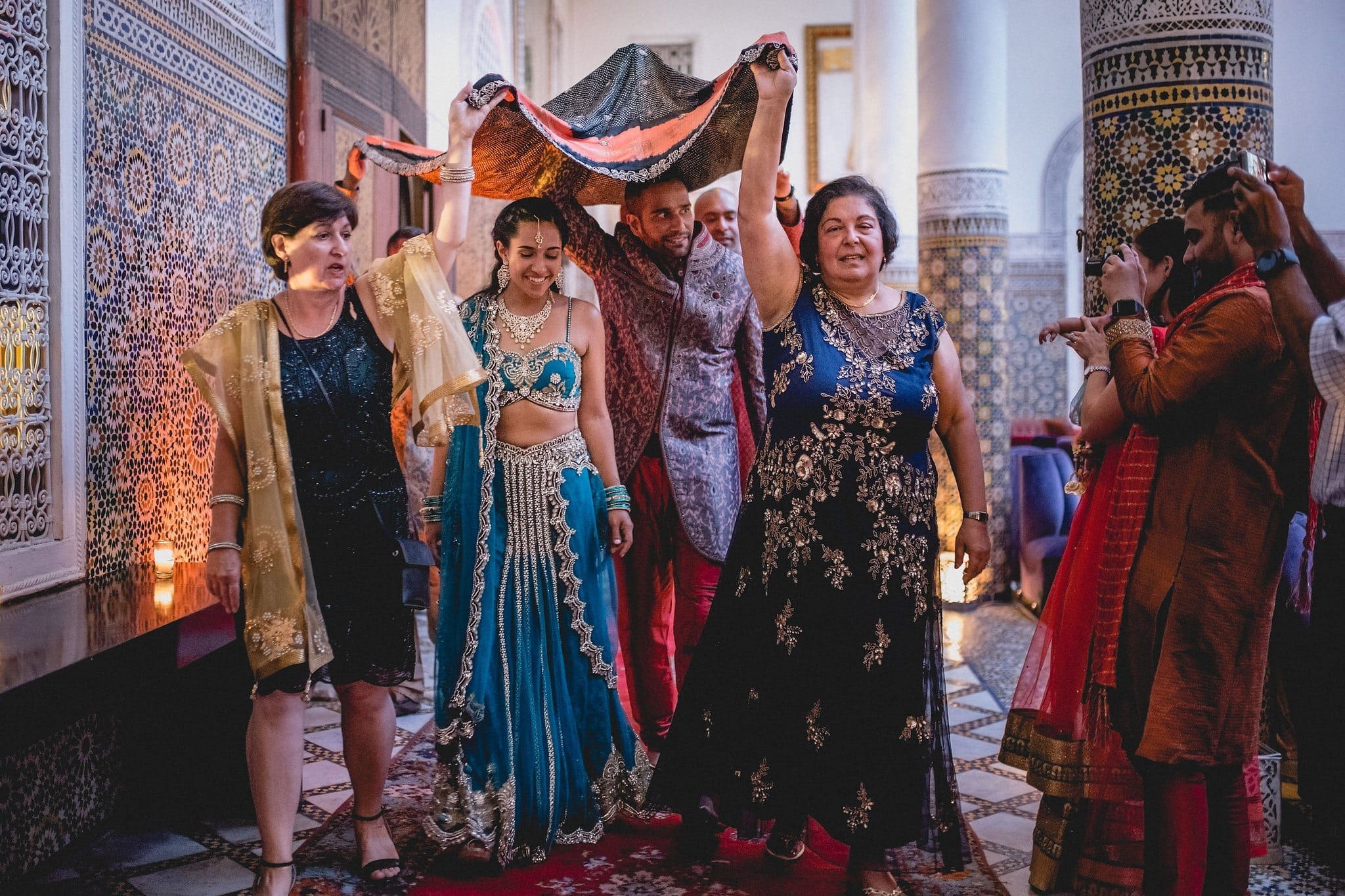 The bride and groom's mothers hold a wedding scarf above the couple as they walk in