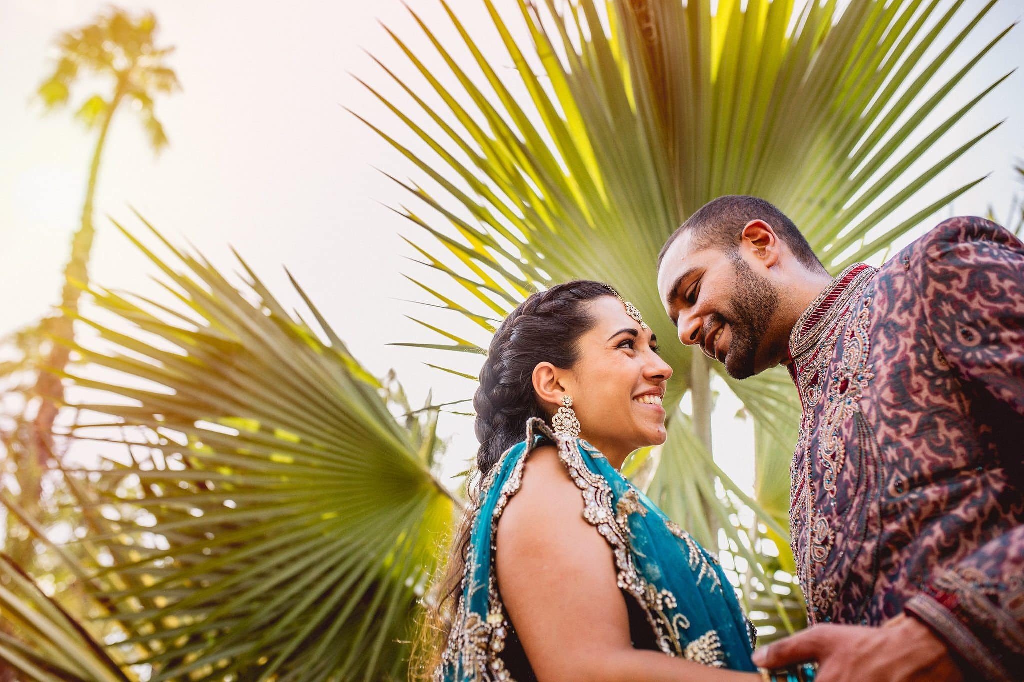 Asian bride and groom smile at each other framed by a large palm tree leaf in Marrakech