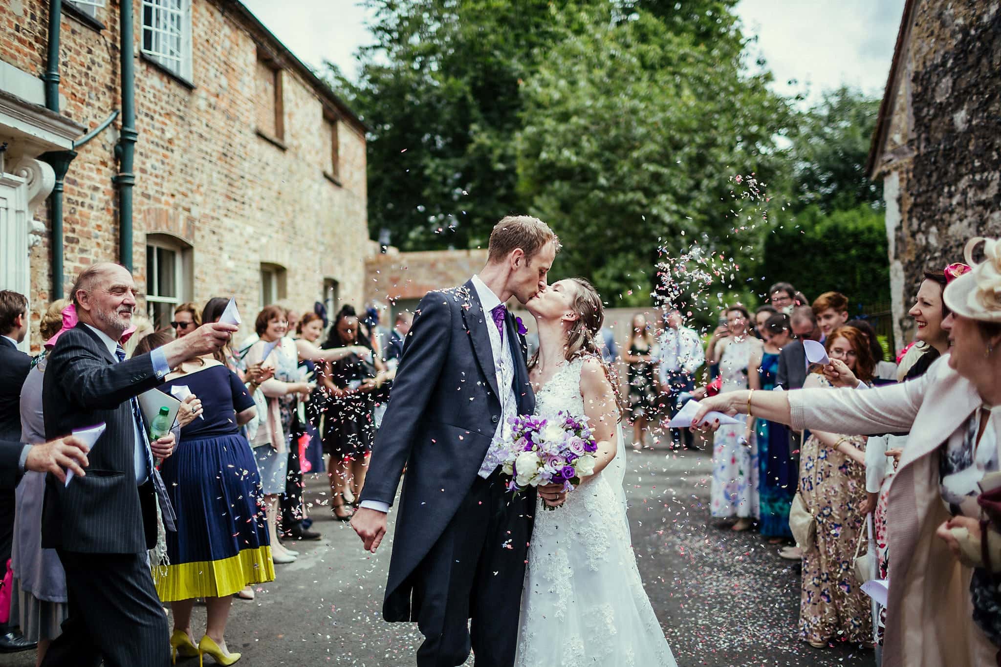 Bride and groom kiss amid purple confetti petals at Dorney Court